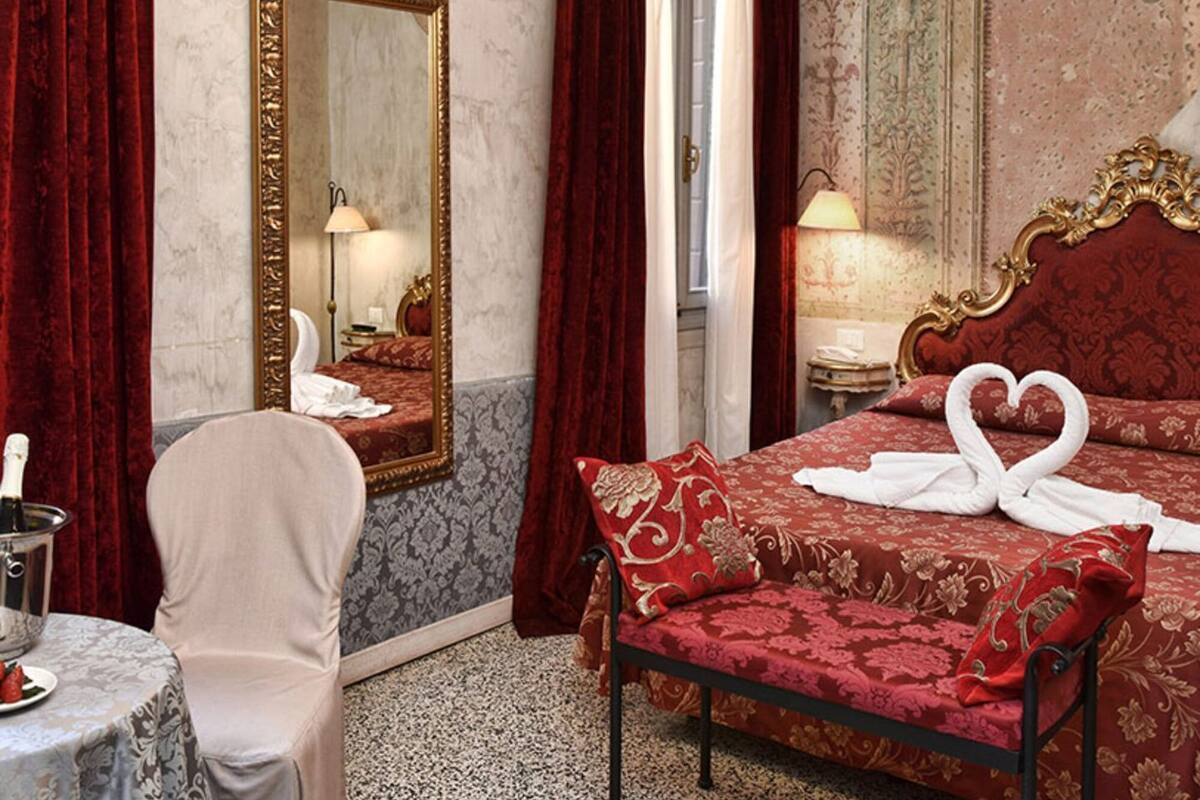Une chambre d’hôtel avec un grand miroir, un lit avec une literie rouge, un lustre et une table sur laquelle se trouvent une bouteille et des verres.