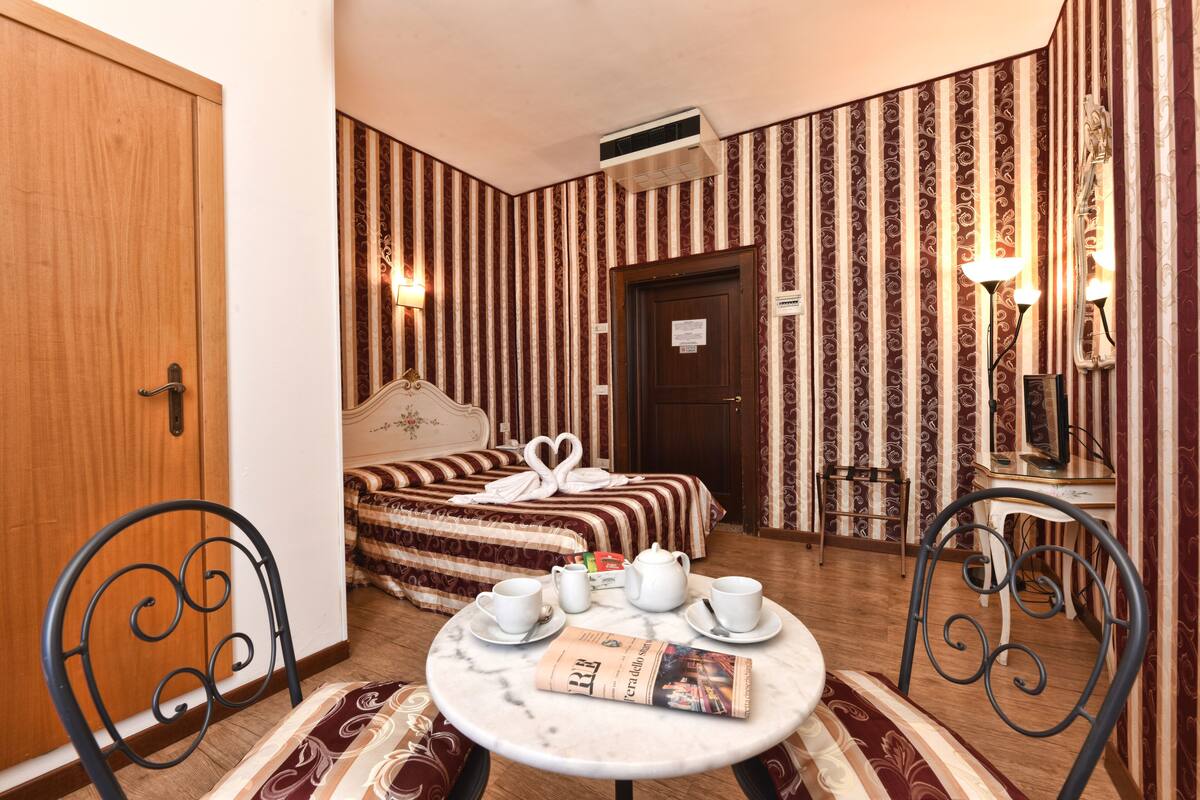 Une chambre d’hôtel avec un lit, une table avec un journal, une chaise, une table de chevet avec une lampe et une armoire.