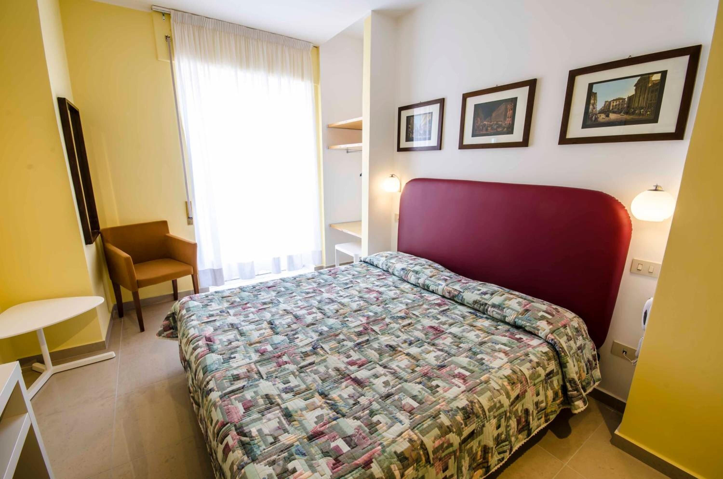A hotel room with a bed, a chair, a small table, and a window with curtains.