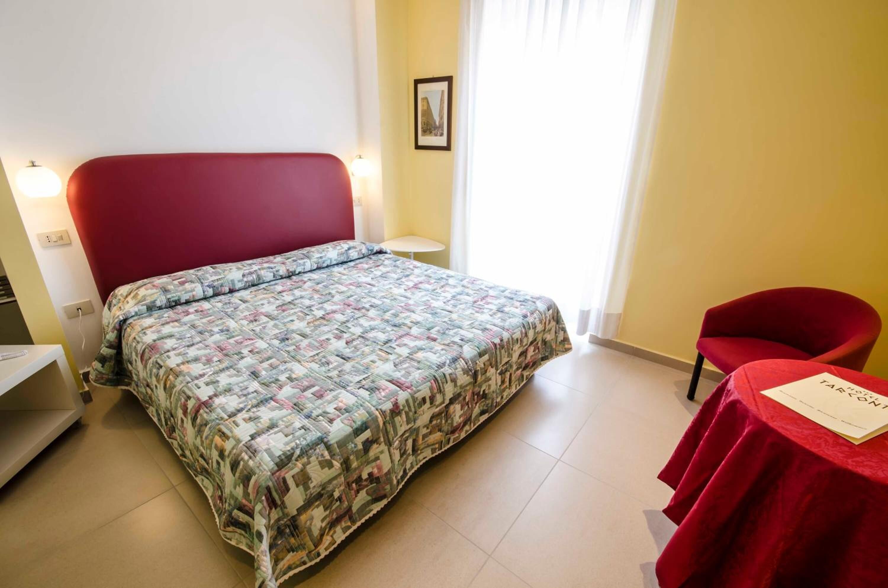 A bedroom with a red headboard, a patterned bedspread, a small bedside table, a chair, and a table with a red tablecloth.