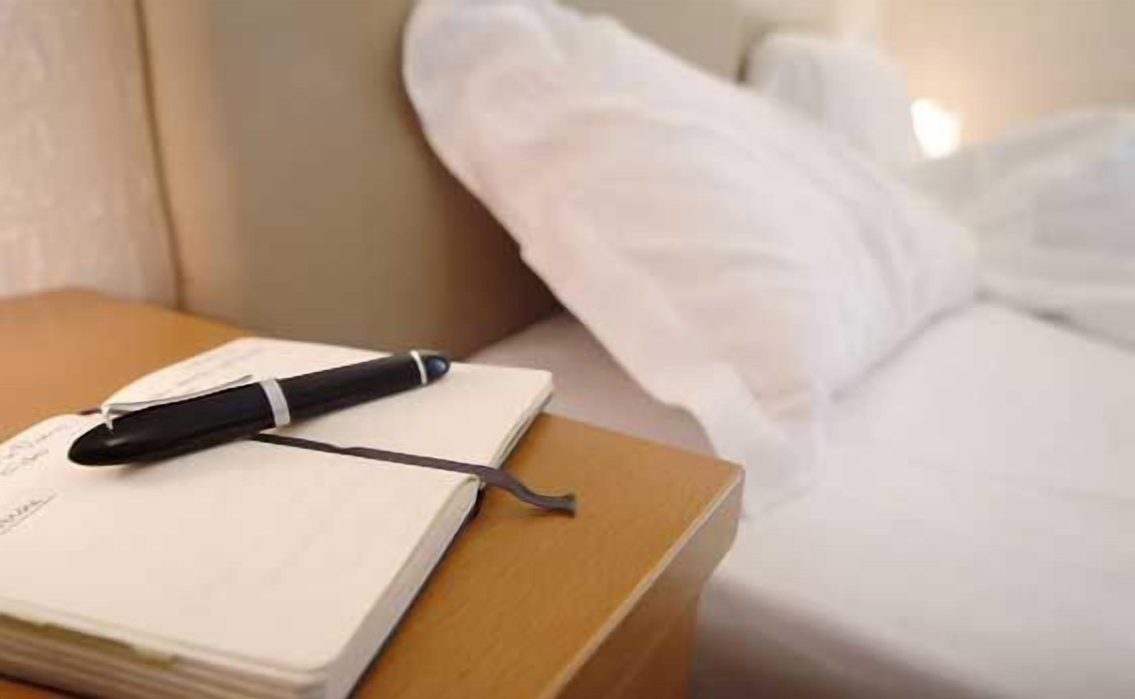 Un cuaderno sobre una mesita de noche de madera con un bolígrafo, junto a una cama con sábanas blancas.