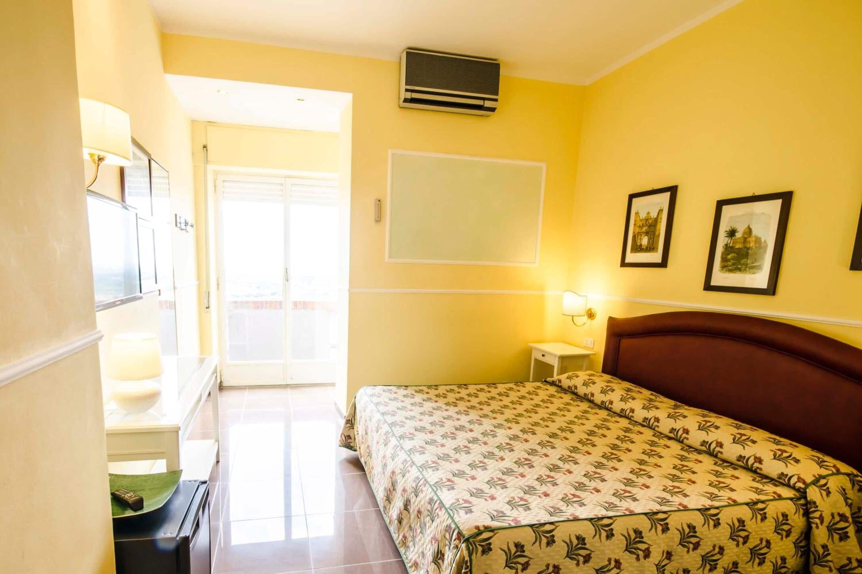 A hotel room with a bed, a desk, a chair, a lamp, a picture frame, and a window.