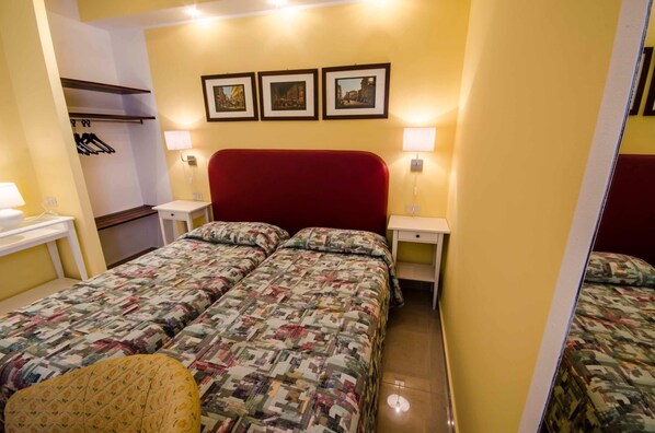 Une chambre d’hôtel avec deux lits, une tête de lit rouge, des lampes de chevet, une petite table et un porte-manteau.
