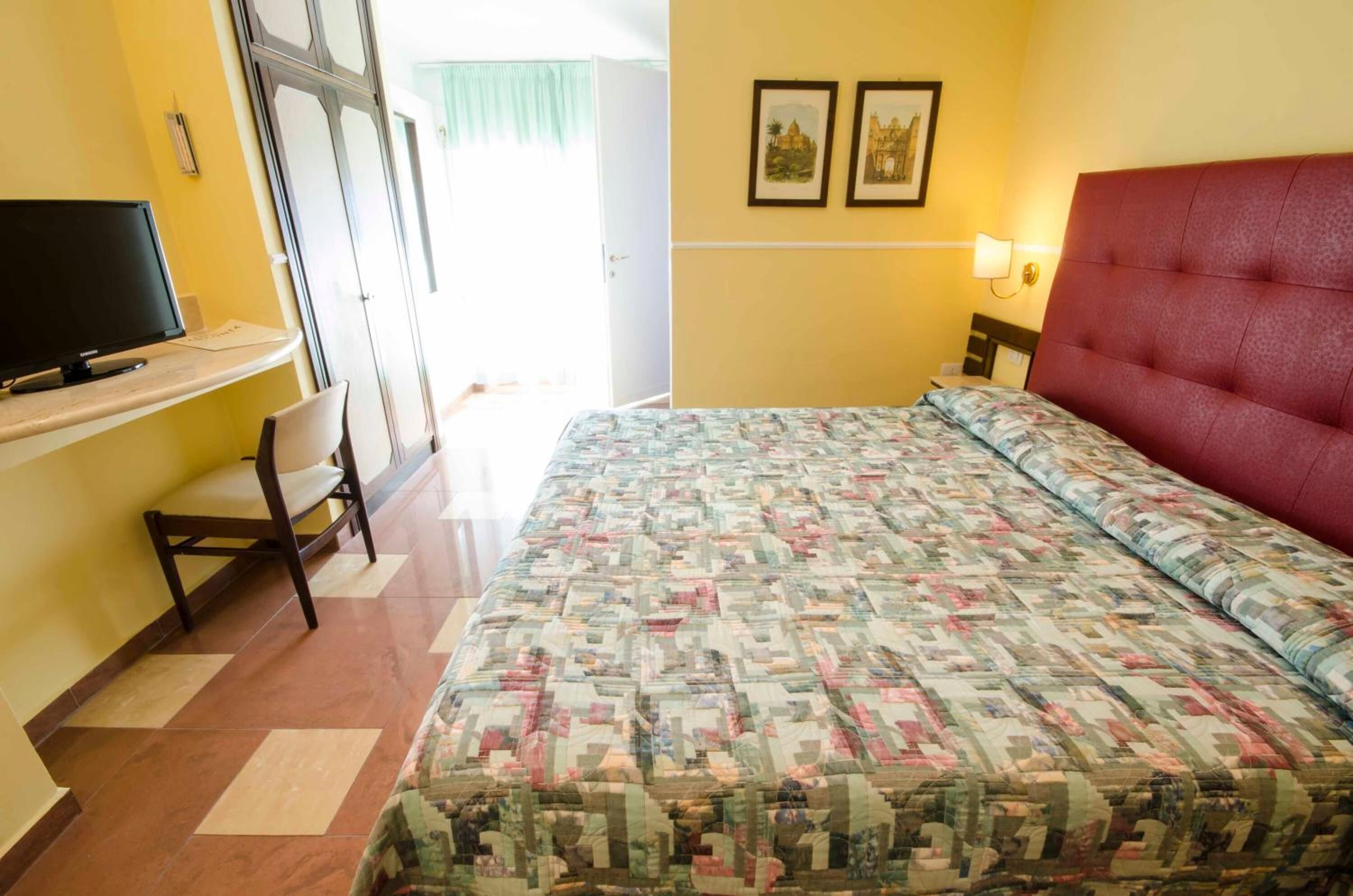 A hotel room with a bed, a desk with a television, a chair, and two framed pictures on the wall.