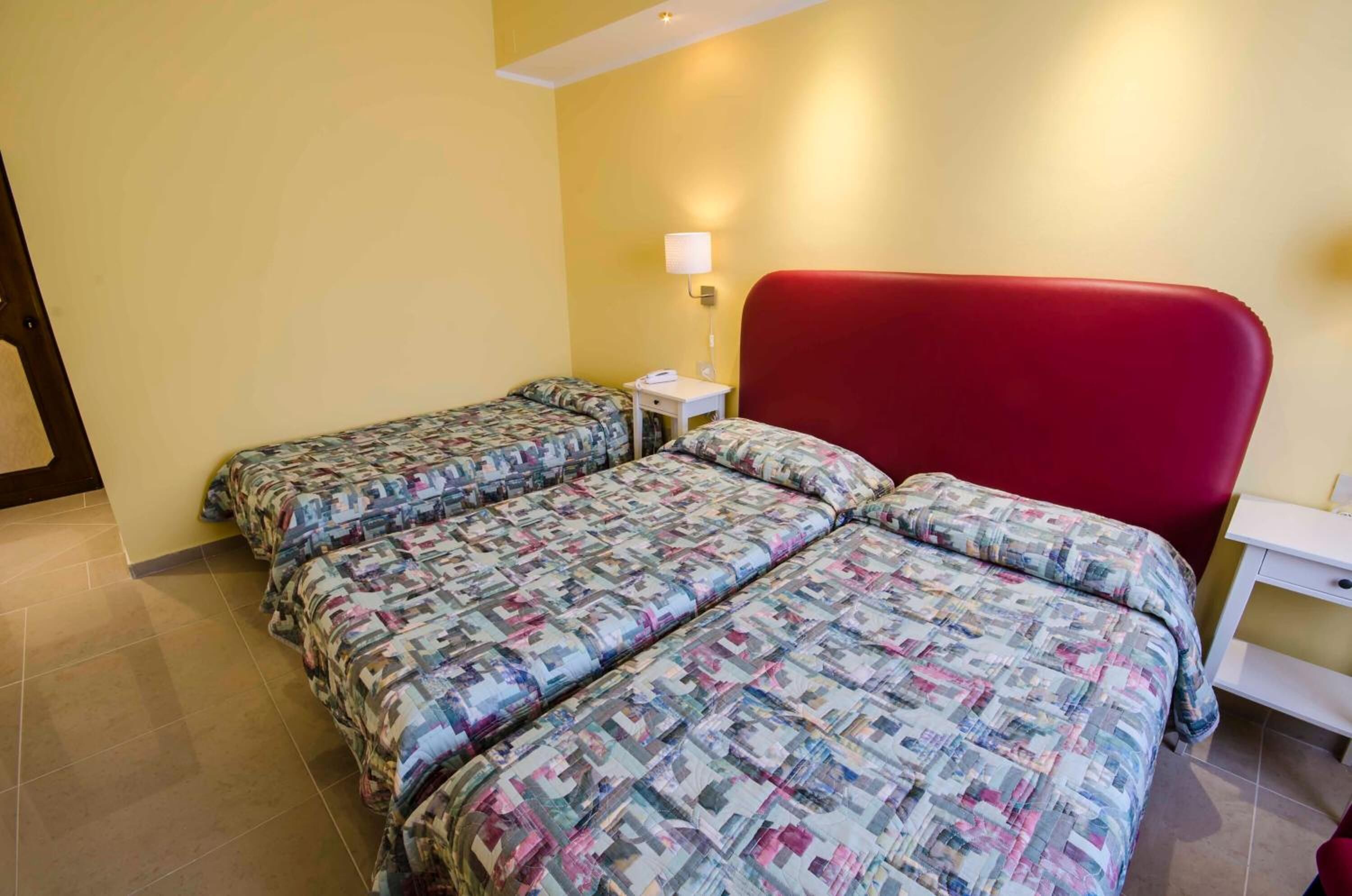 Una habitación con dos camas, cabecera roja, mesita de noche con lámpara y cómoda blanca.