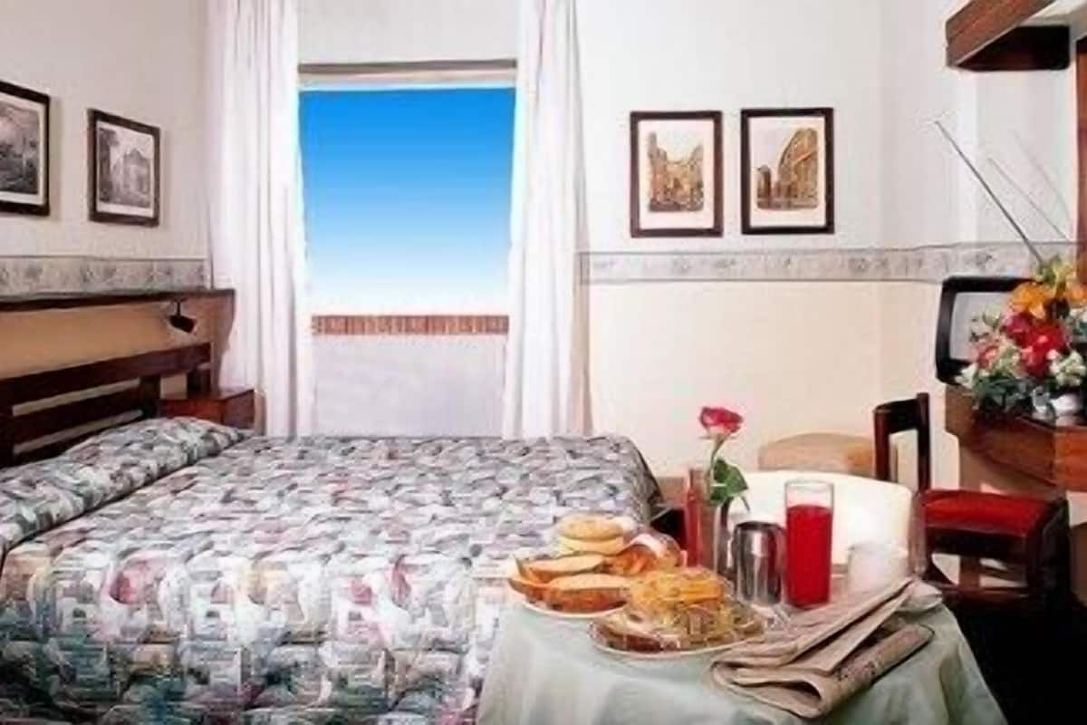 A hotel room with a bed, a table with breakfast items, a window with a view, and framed pictures on the wall.
