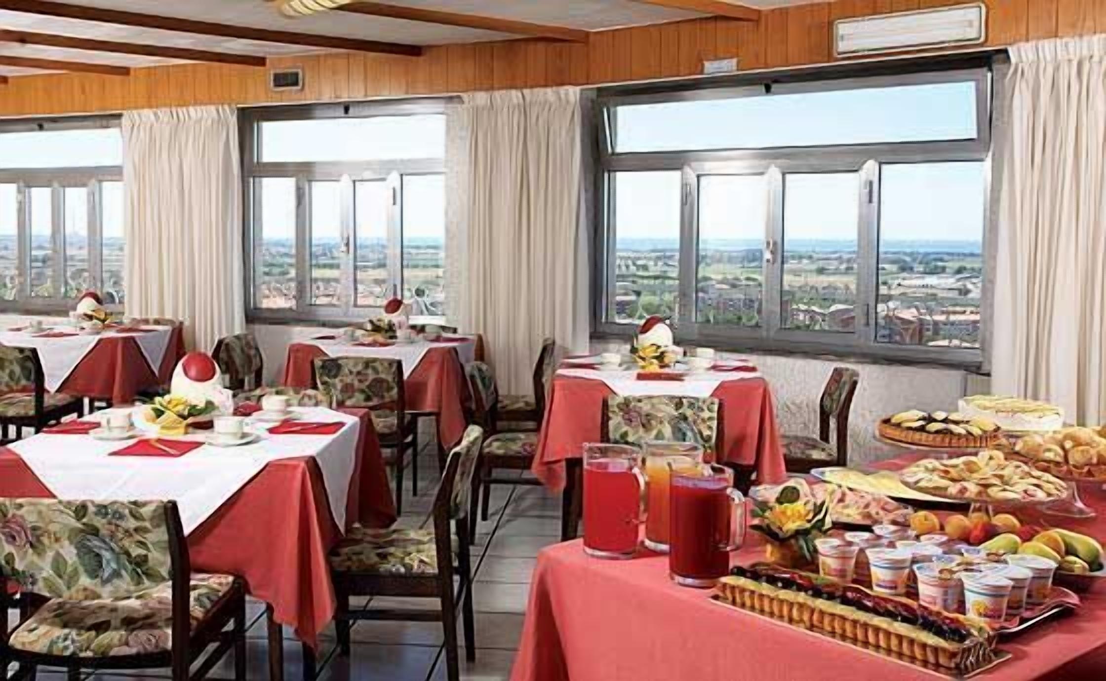 A dining area with tables set for breakfast, featuring a variety of fruits and pastries, and large windows offering a view of the countryside.
