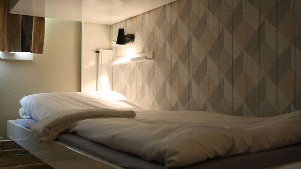 A neatly made bed with a white comforter and pillows, a bedside lamp, and a wall with a geometric pattern. - HI Oslo Haraldsheim - Hostel (Oslo)