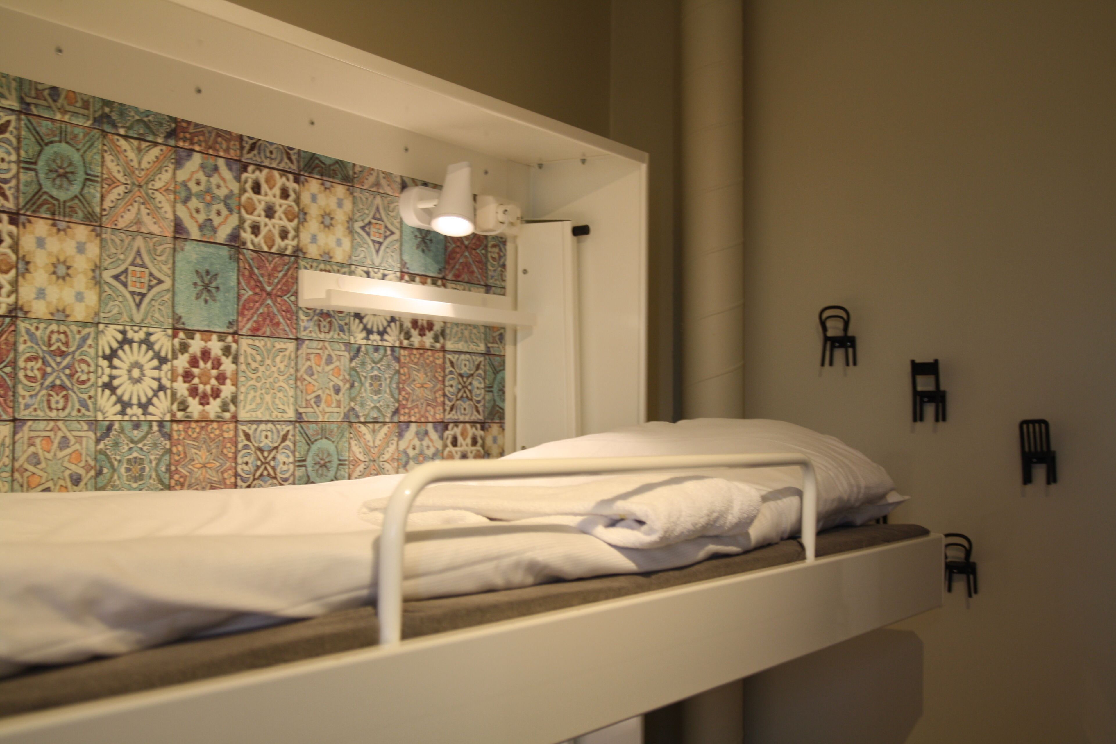 Una cama litera con una pared de azulejos detrás, un pequeño escritorio con una lámpara y una silla junto a la pared.