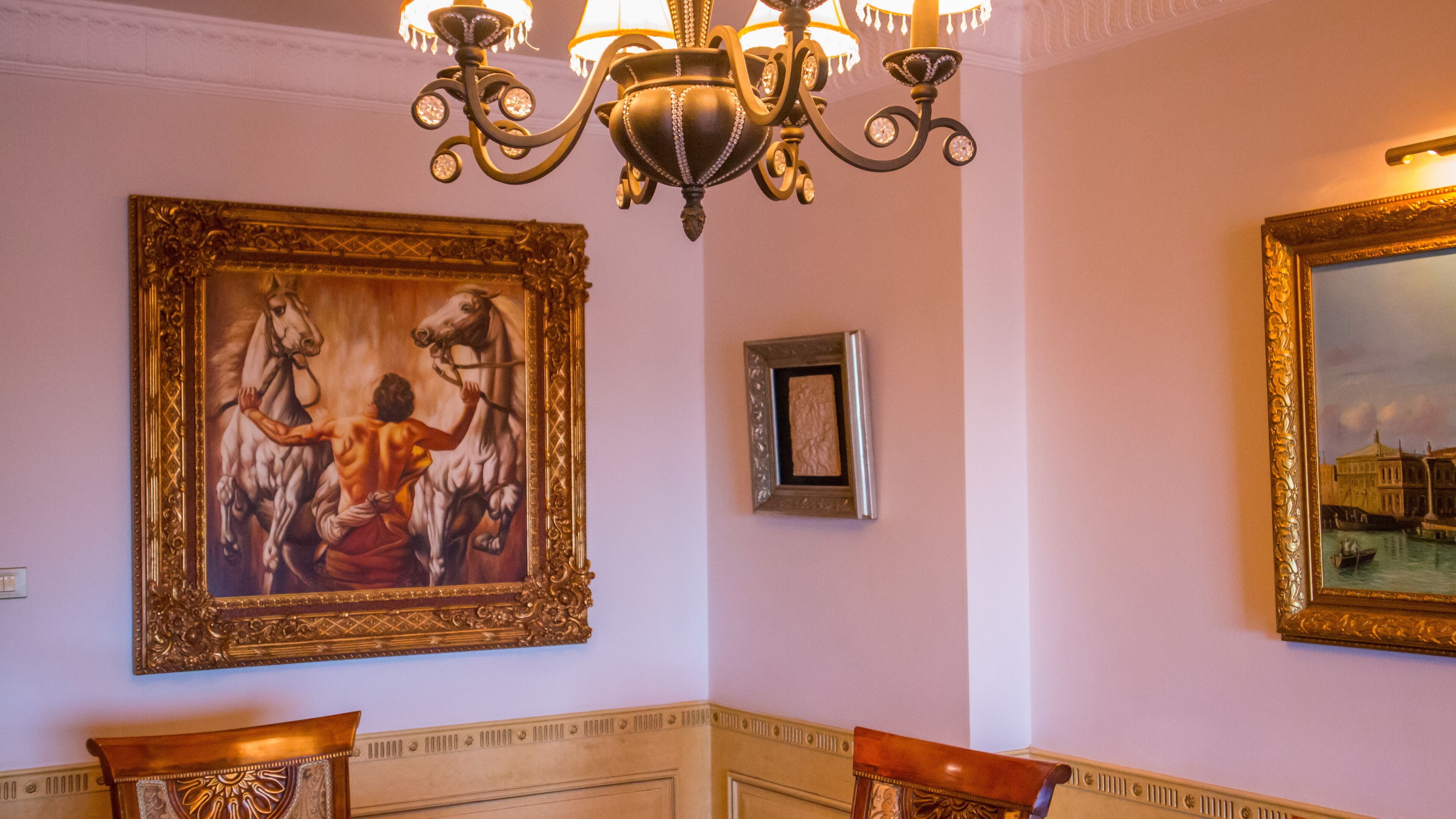 A room with a large framed painting of a classical scene, a smaller framed picture, and a chandelier.