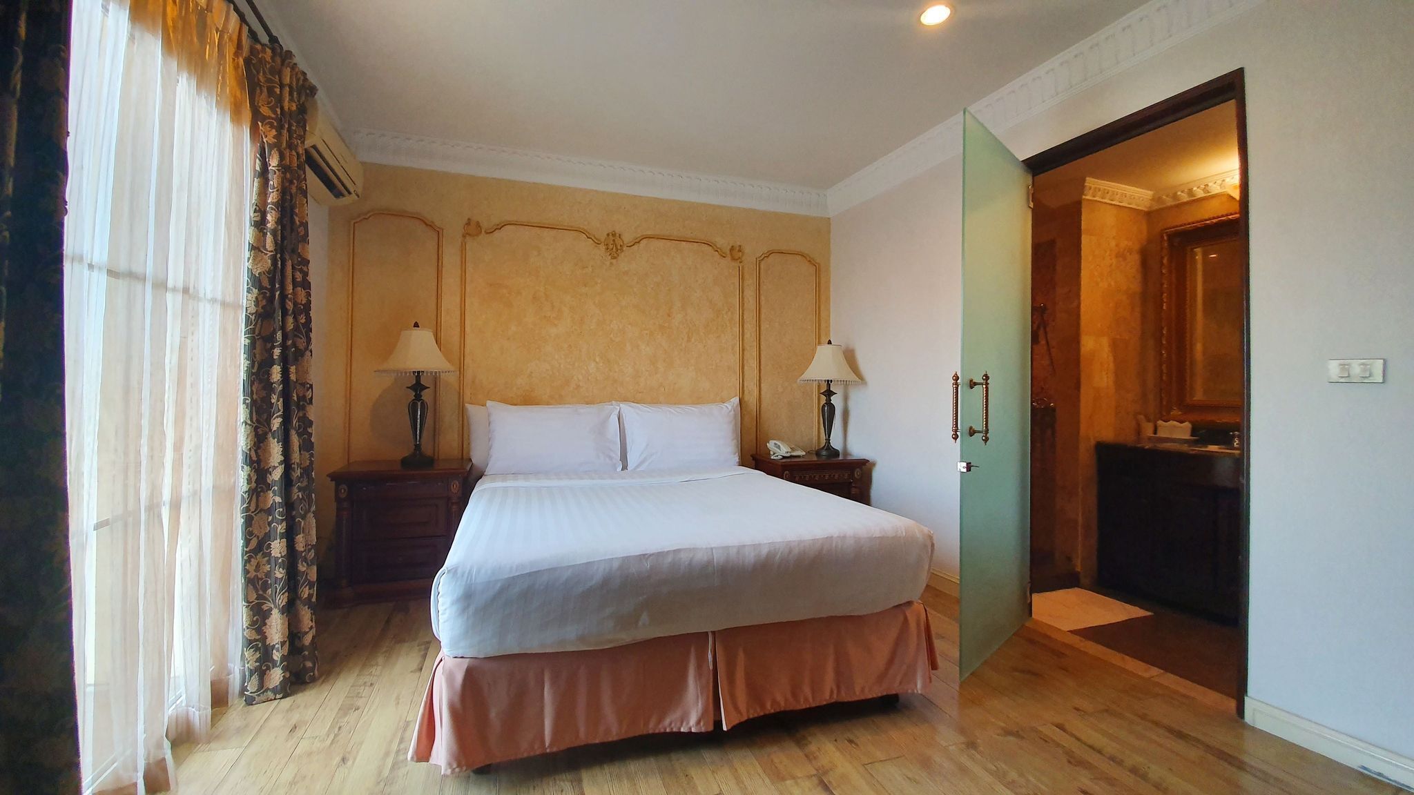A hotel room with a large bed, two bedside tables with lamps, a wooden floor, and a green door leading to a bathroom.