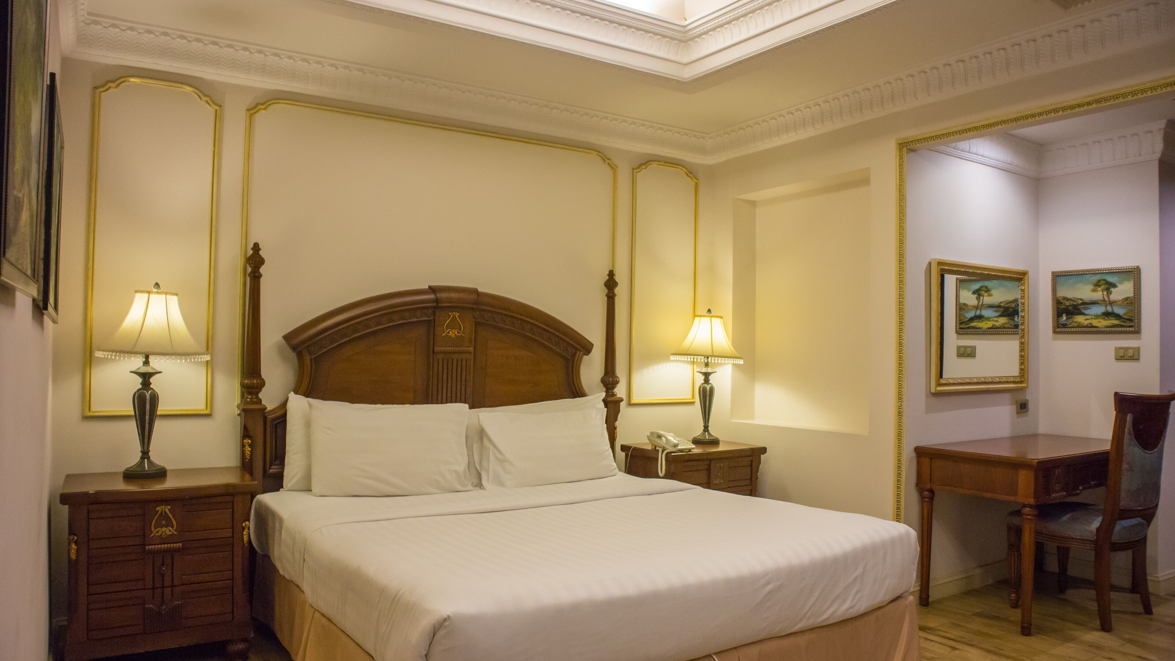 A hotel room with a large bed, two bedside tables with lamps, a wooden desk, and chairs.