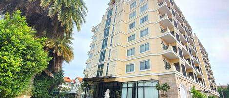 A multi-story hotel with a beige facade, balconies on each floor, and a landscaped entrance area with potted plants.