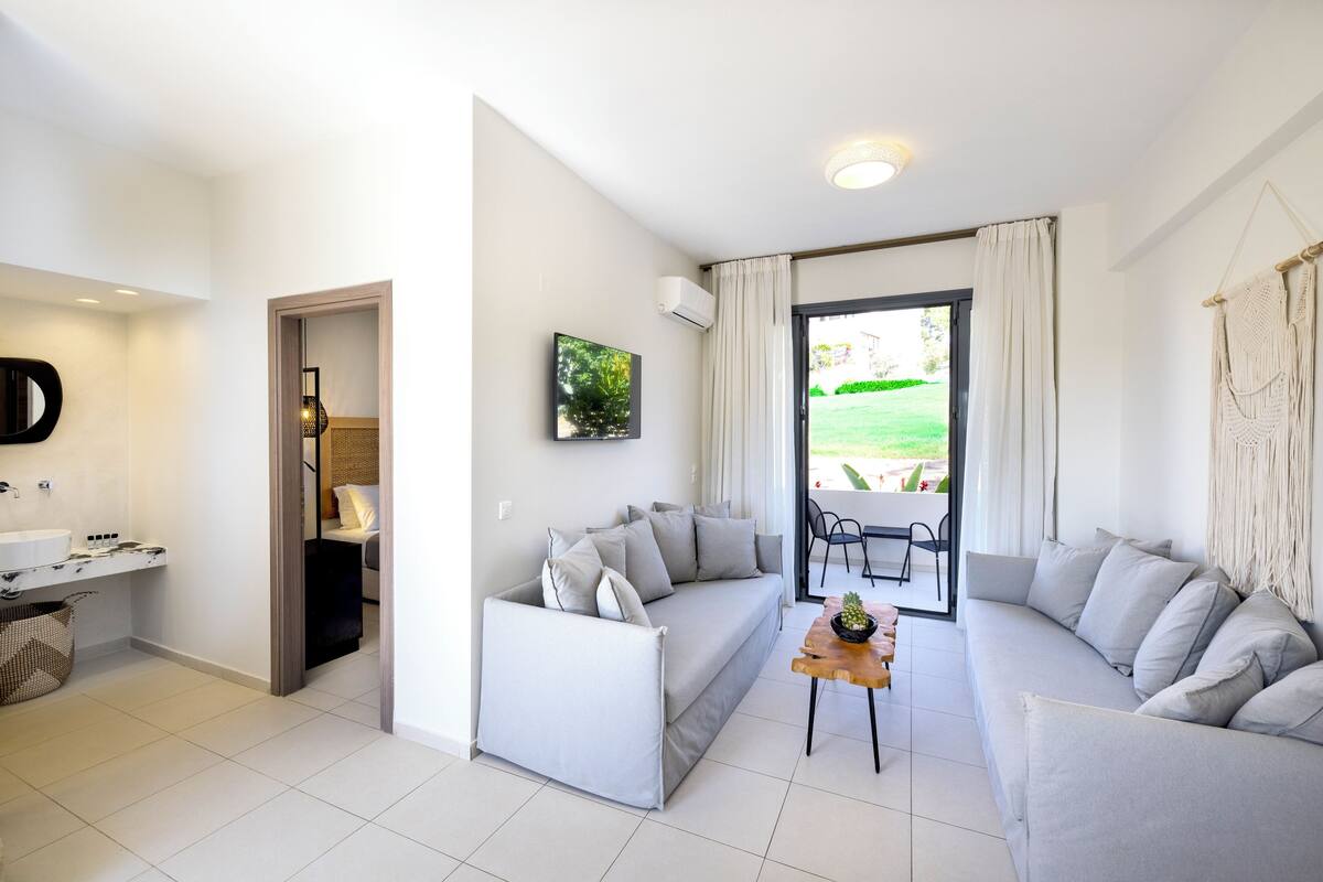 Sala de estar moderna com um sofá cinzento, uma mesa de centro de madeira e vista para um jardim através de uma porta de vidro.