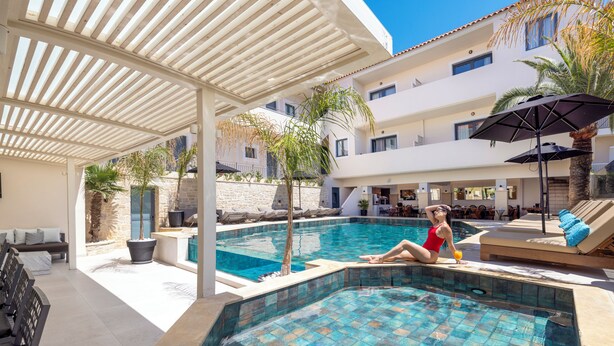 A pool area with a person relaxing by the pool, lounge chairs, and umbrellas. - La Stella Suites (Rethymno)