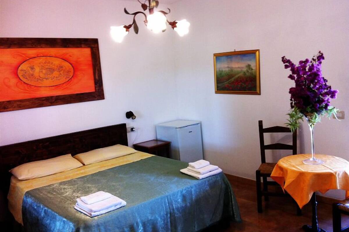A hotel room with a bed, a desk, a chair, a small table with a vase of flowers, and two framed pictures on the wall.