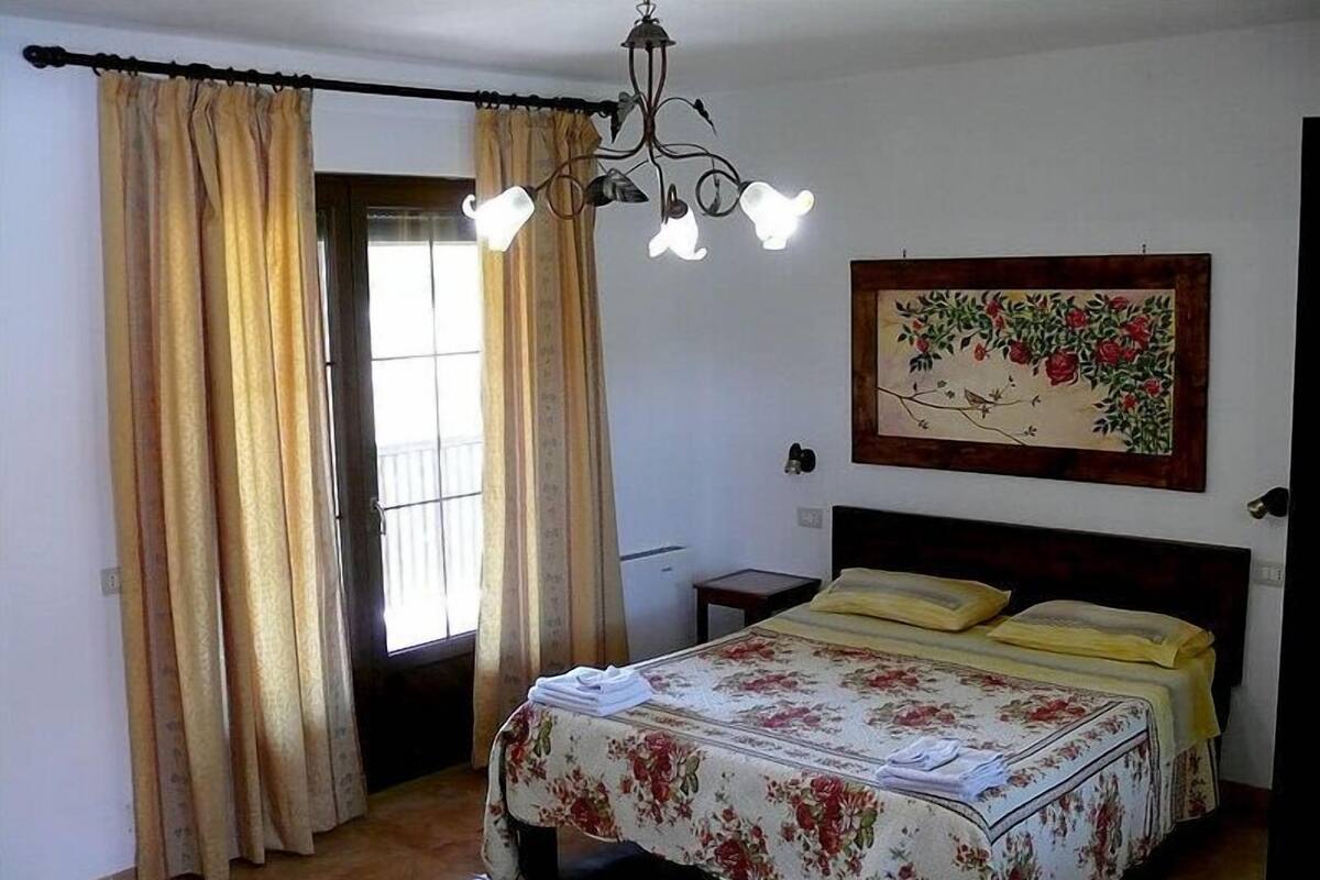 A bedroom with a bed, curtains, a chandelier, a nightstand, and a floral painting on the wall.