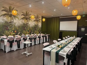 Un salón de conferencias con mesas dispuestas para una reunión, una pantalla de proyector y un mural tropical en la pared.