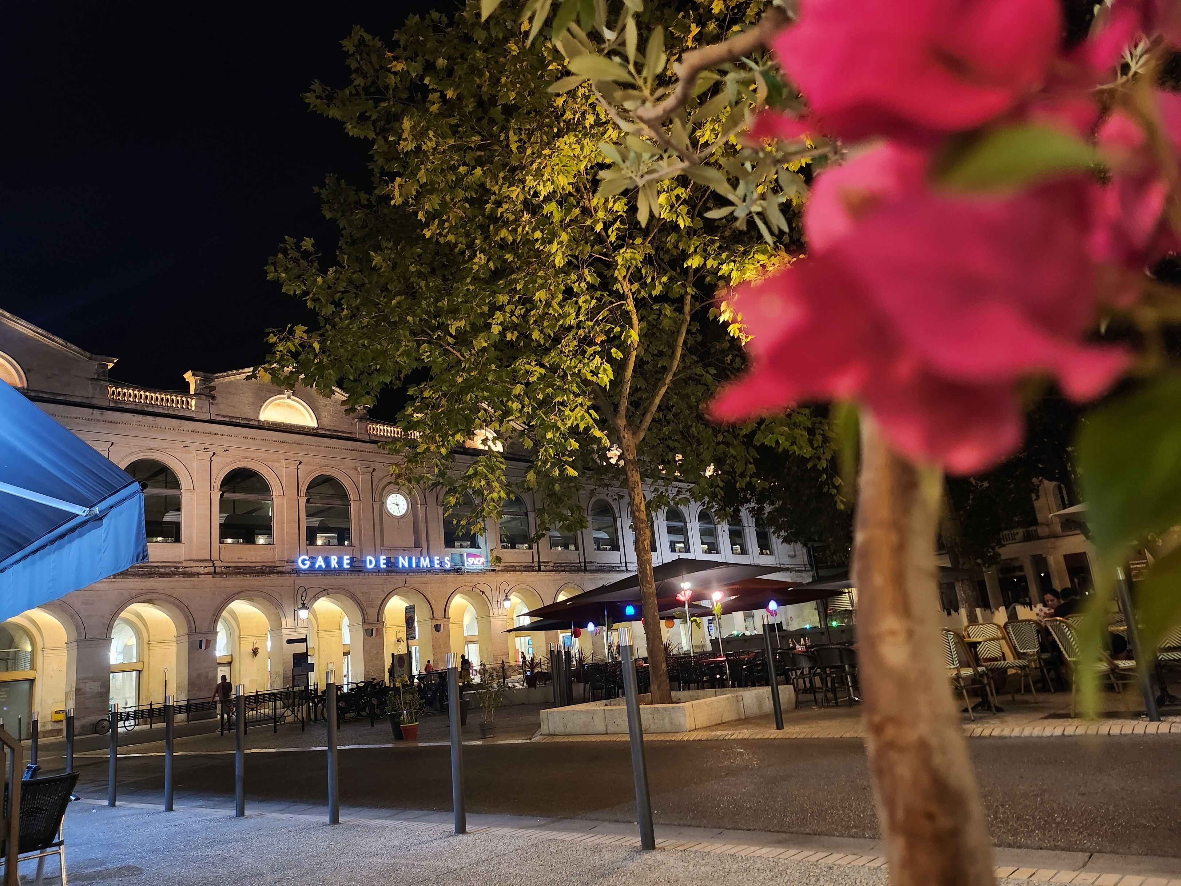 A building with a sign reading 'GARE DE NIMES' and a covered outdoor seating area with tables and chairs.
