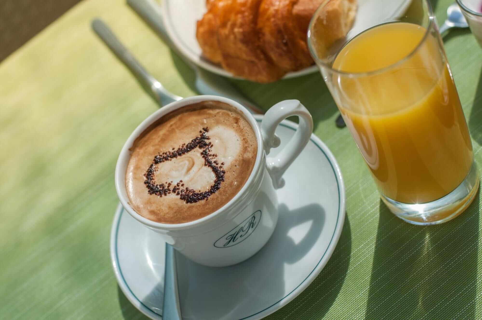 Una taza de café con arte de latte sobre un platito, un vaso de zumo de naranja y un croissant en un plato.
