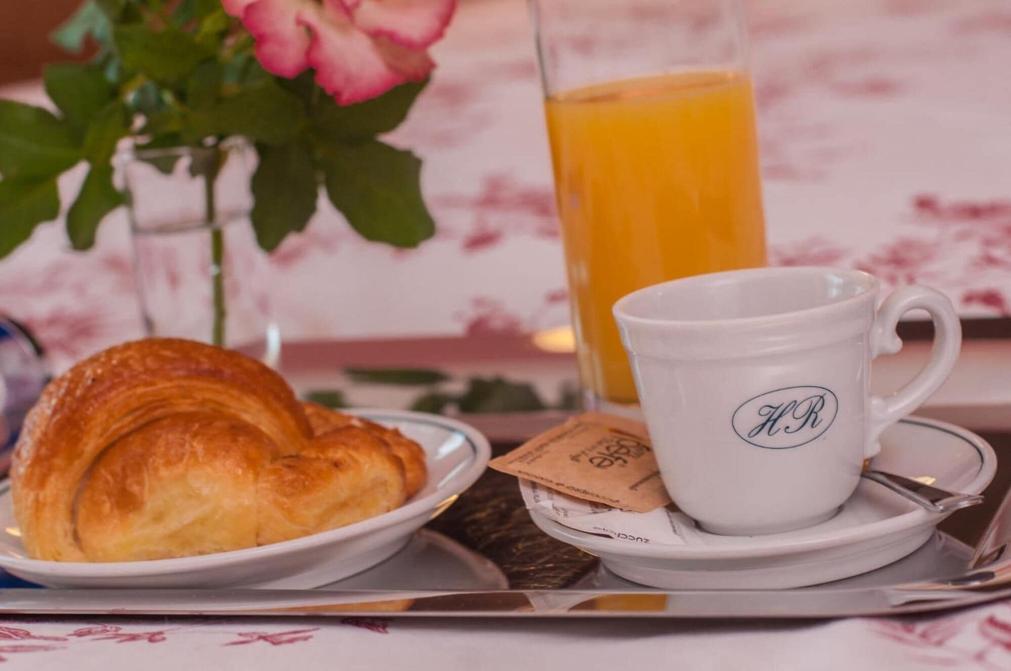 Un desayuno con un croissant, una taza blanca con iniciales y un vaso de jugo de naranja sobre una mesa.