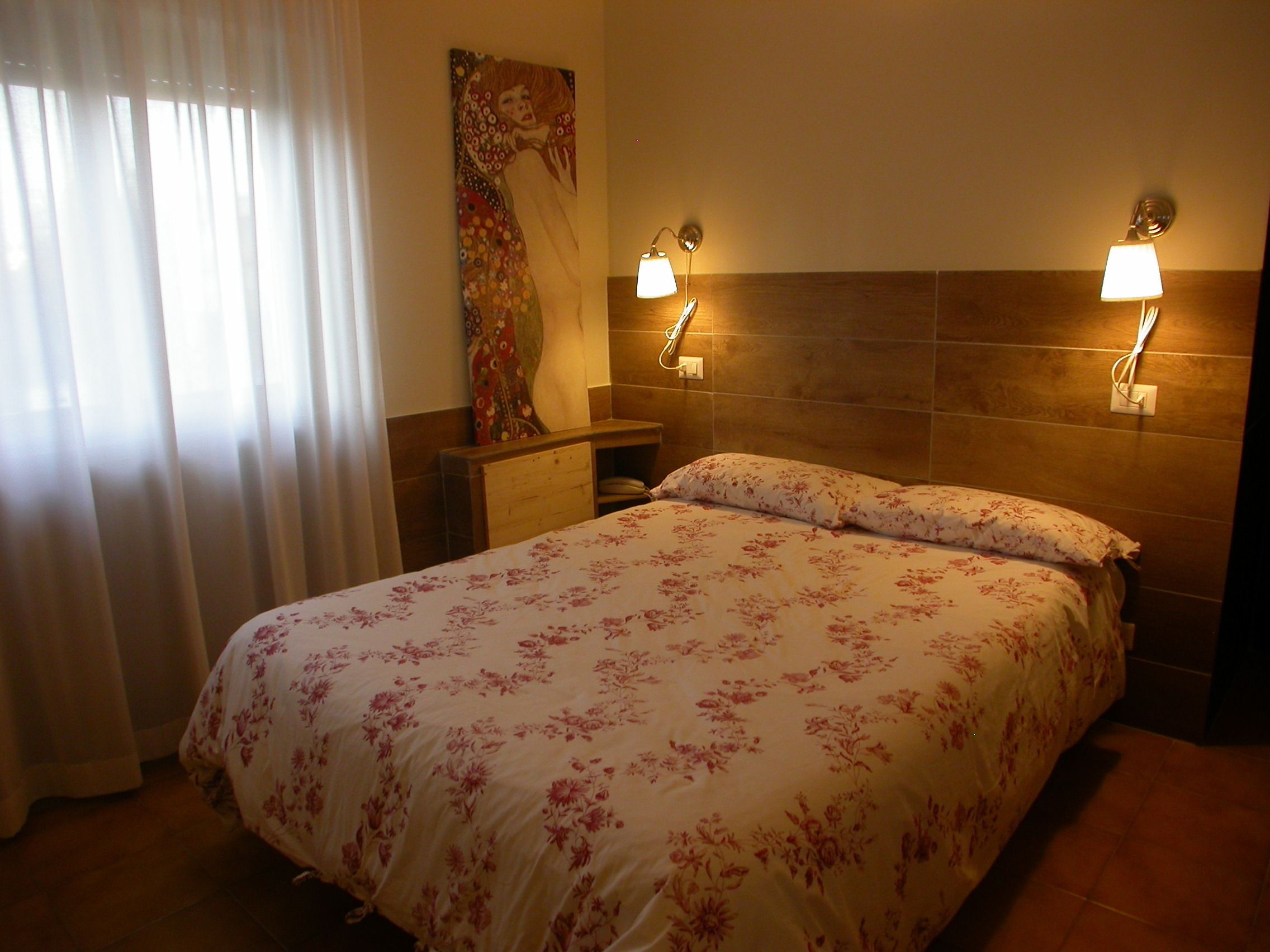 A hotel room with a bed, two wall-mounted lamps, a painting on the wall, and a window with curtains.