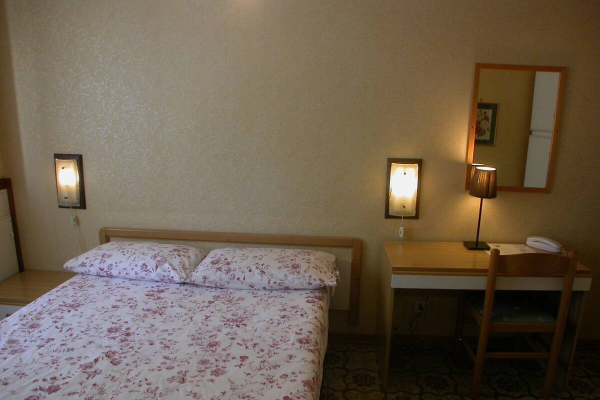 A hotel room with a bed, a desk, and a chair. There are two wall-mounted lamps and a mirror on the wall.