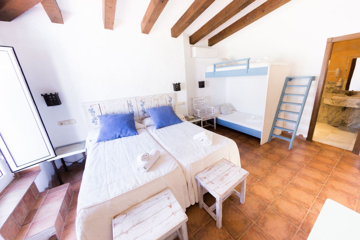 Une chambre avec deux lits, un banc en bois, une chaise et une salle de bain visible à travers une porte ouverte.