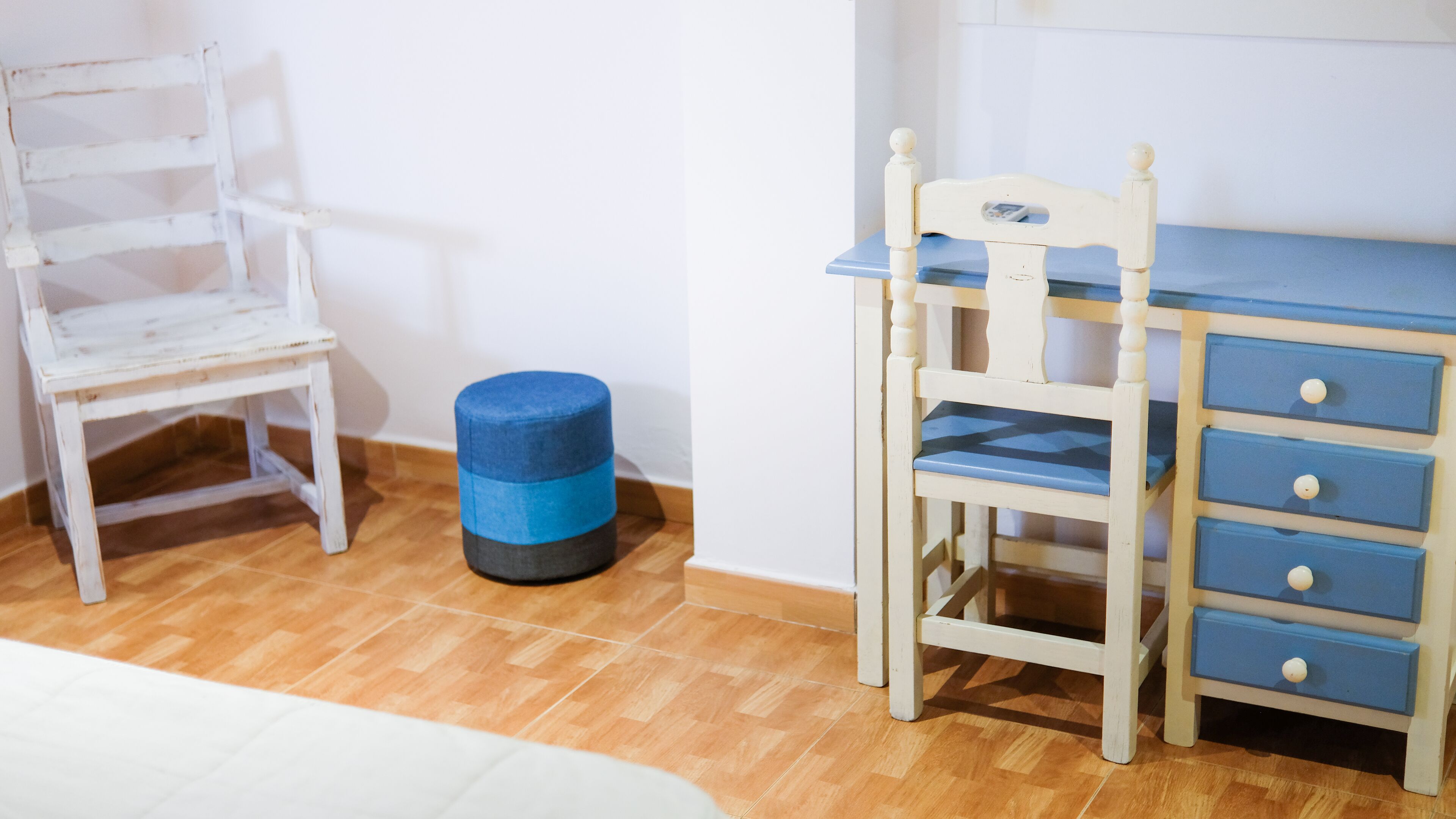 Una habitación con piso de madera, una silla blanca, un taburete azul y un escritorio azul con cajones.