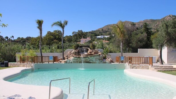 A swimming pool with a waterfall feature, surrounded by palm trees and a hilly backdrop. - La Torre (Tarifa)
