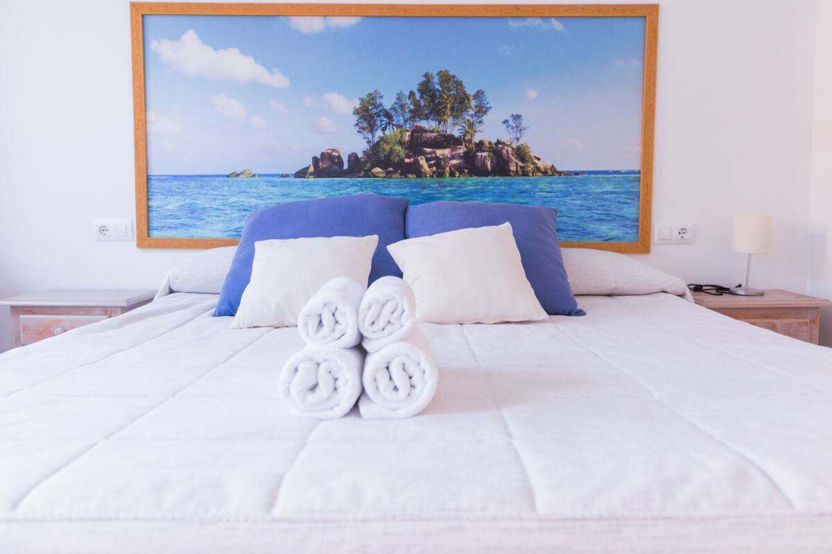 Un lit bien fait, avec du linge de lit blanc et des serviettes pliées, un grand tableau représentant une île tropicale accroché au mur, et une table de chevet avec une lampe.