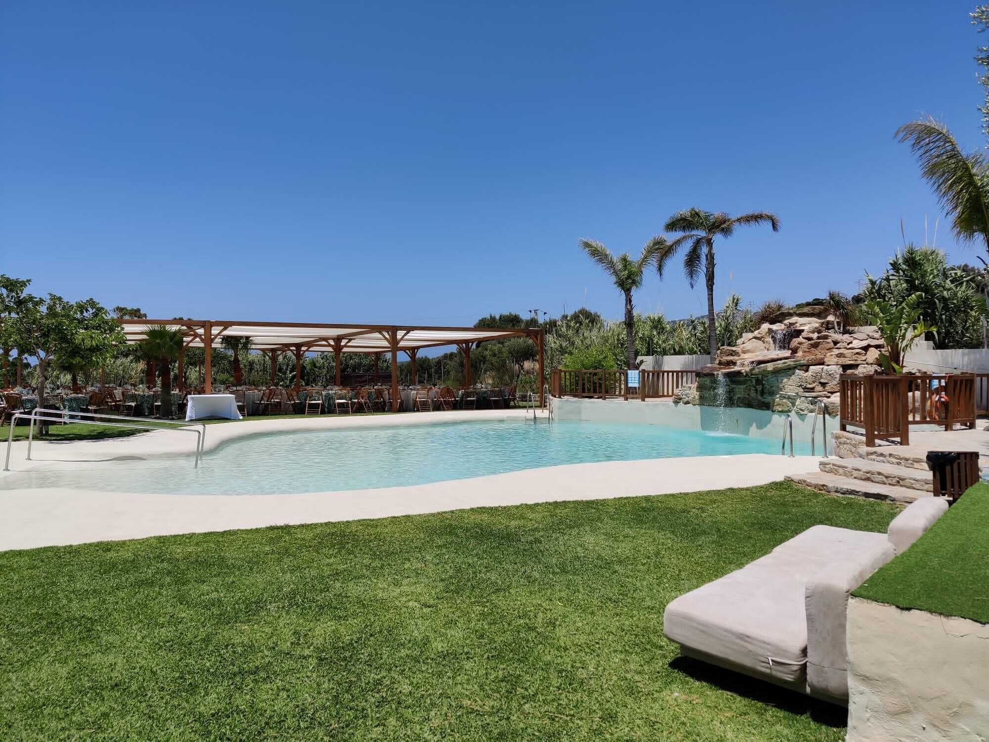 A swimming pool with a pergola and lounge chairs, surrounded by palm trees and a grassy area.