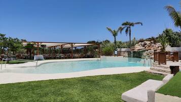 A swimming pool with a pergola and lounge chairs, surrounded by palm trees and a grassy area.