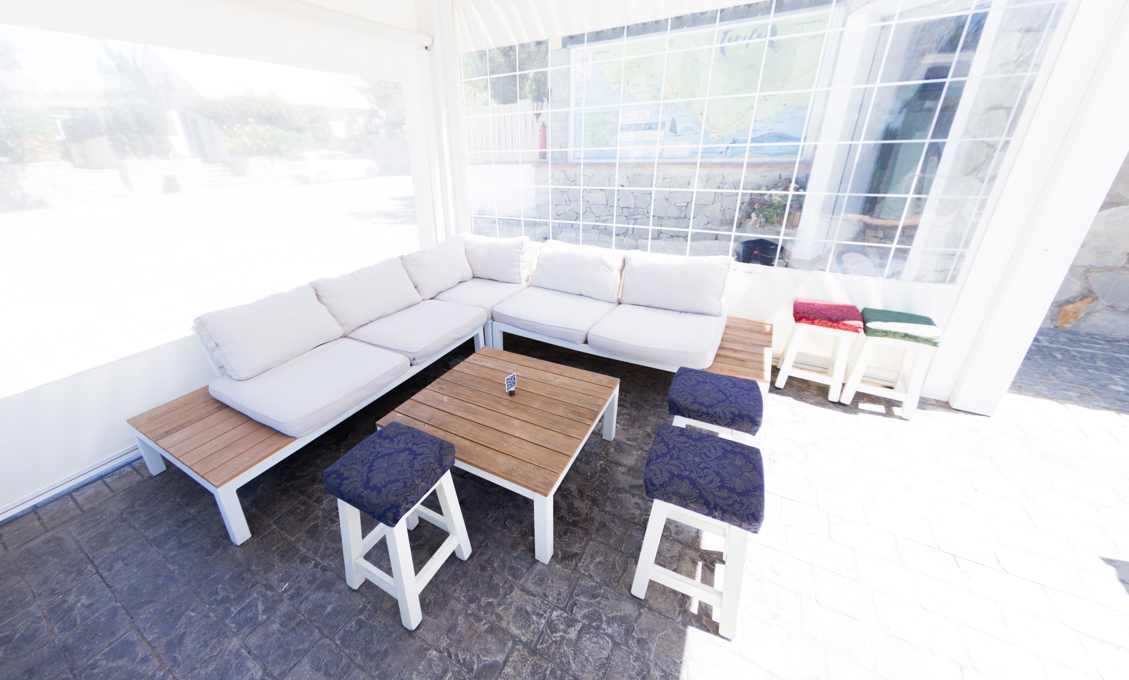 Un espacio al aire libre moderno con un sofá seccional blanco, dos mesas de madera y dos taburetes con cojines azules.