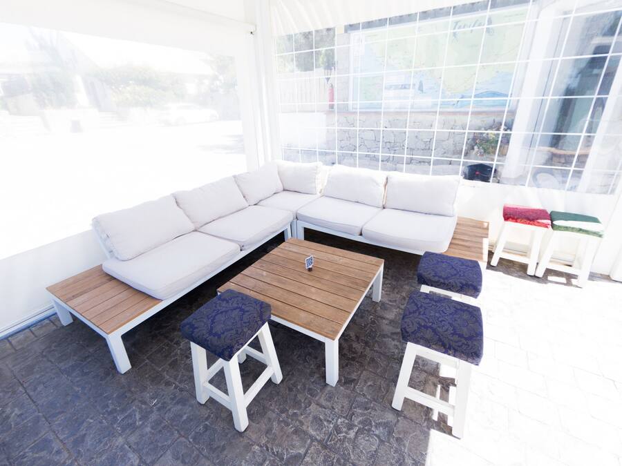 Un espacio al aire libre moderno con un sofá seccional blanco, dos mesas de madera y dos taburetes con cojines azules.