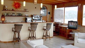 Una zona de bar con tres taburetes de mimbre, una cafetera y estantes con botellas.