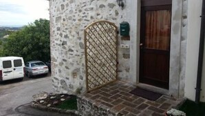 A stone building with a wooden door and a lattice gate. There are parked cars and a green plant in front. - Modà Antica Dimora (Montegiardino)