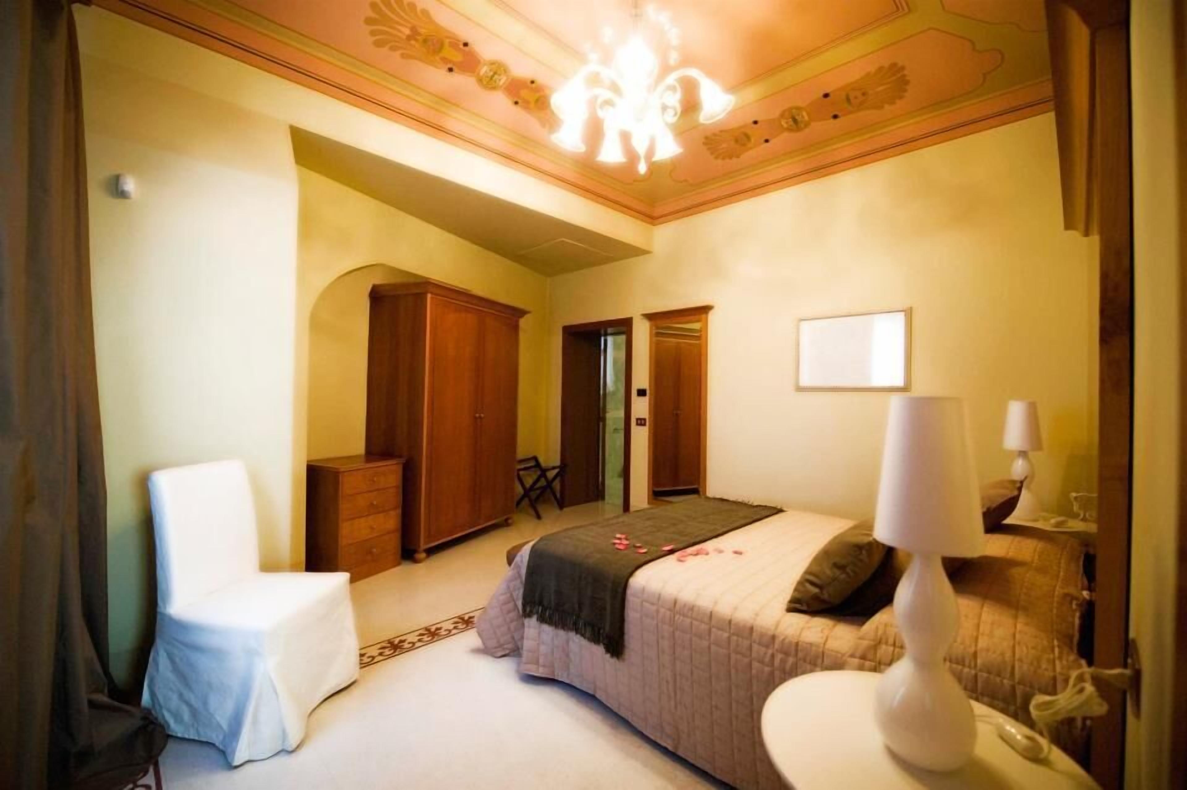 A bedroom with a bed, a chair, a desk, a wardrobe, and a chandelier.