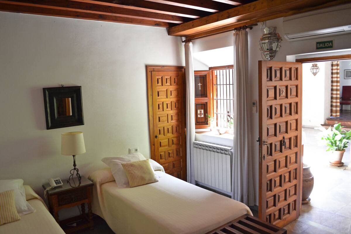A hotel room with two beds, a nightstand with a lamp, a wooden door, and a view of the outside.