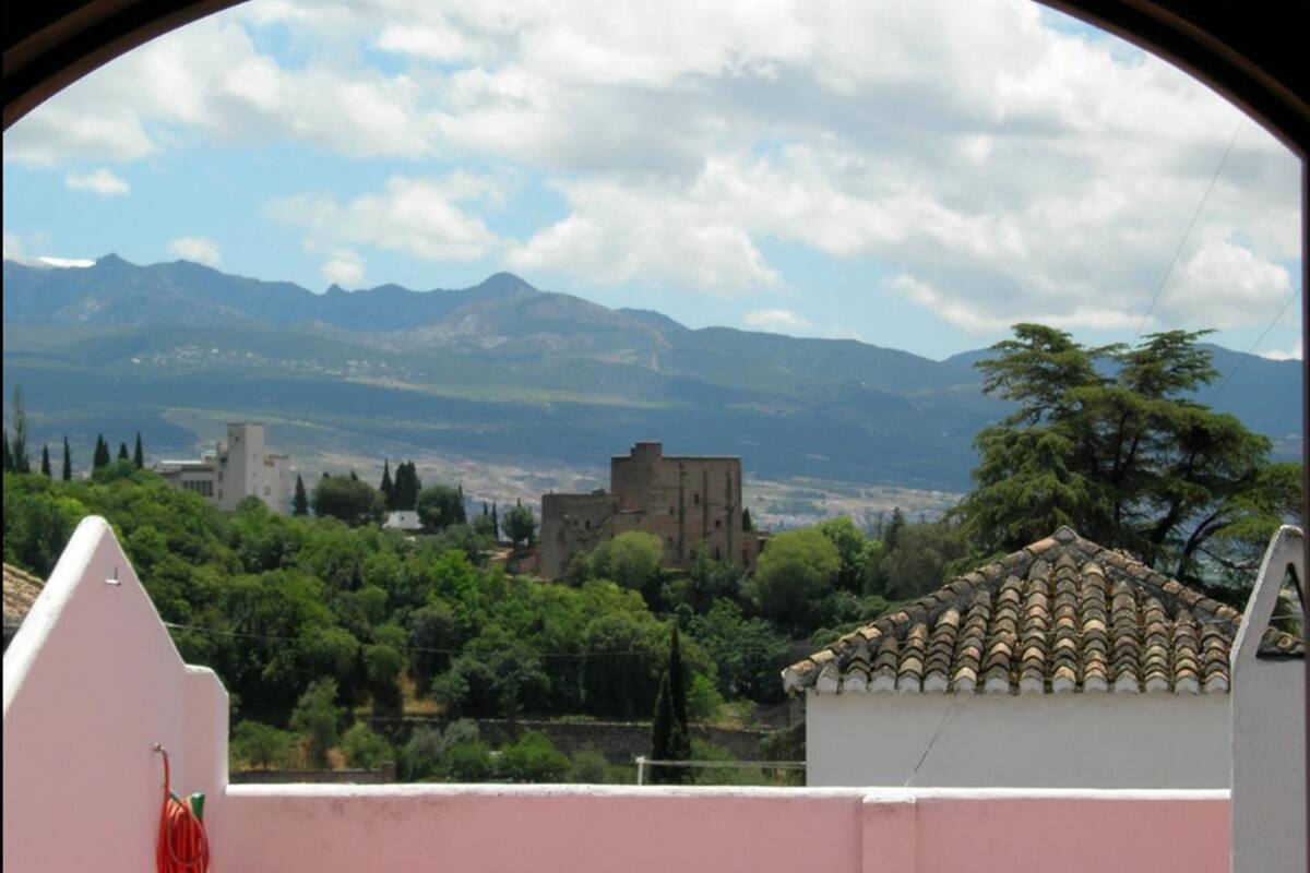 A view through a window of a building with a tiled roof, a mountainous landscape, and a few trees in the foreground.