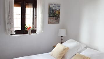A neatly made bed with white linens and striped pillows, a bedside lamp, a window with curtains, and a painting on the wall.