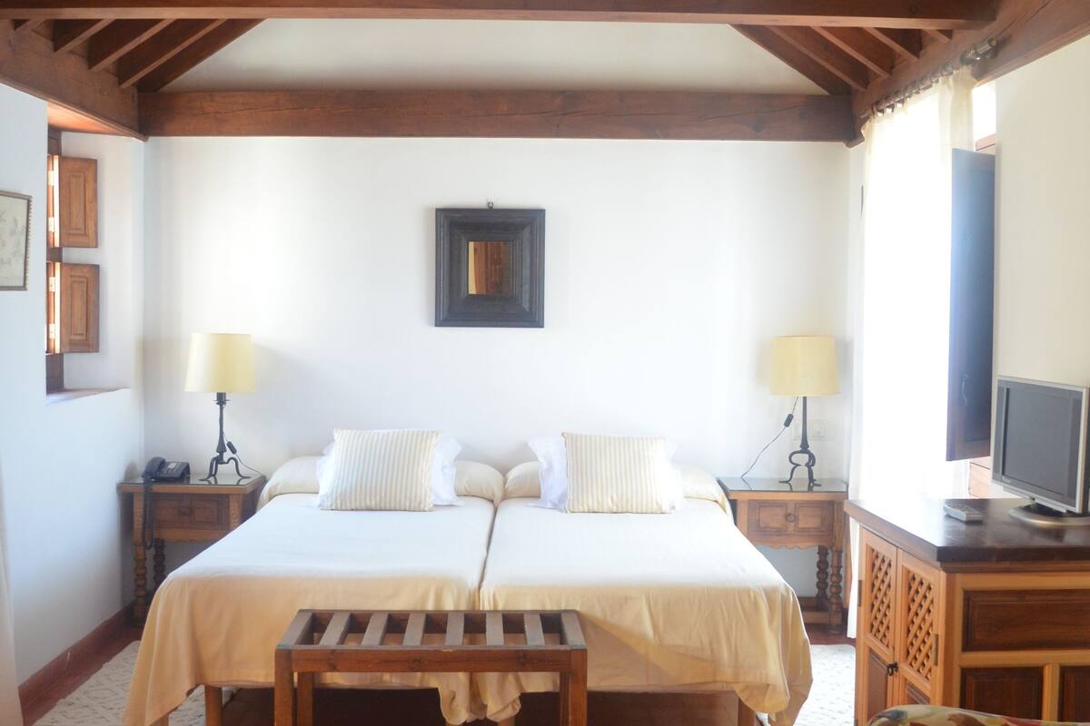A bedroom with a large bed, two bedside tables, a round glass table, a sofa, wooden beams on the ceiling, and a television on a wooden cabinet.