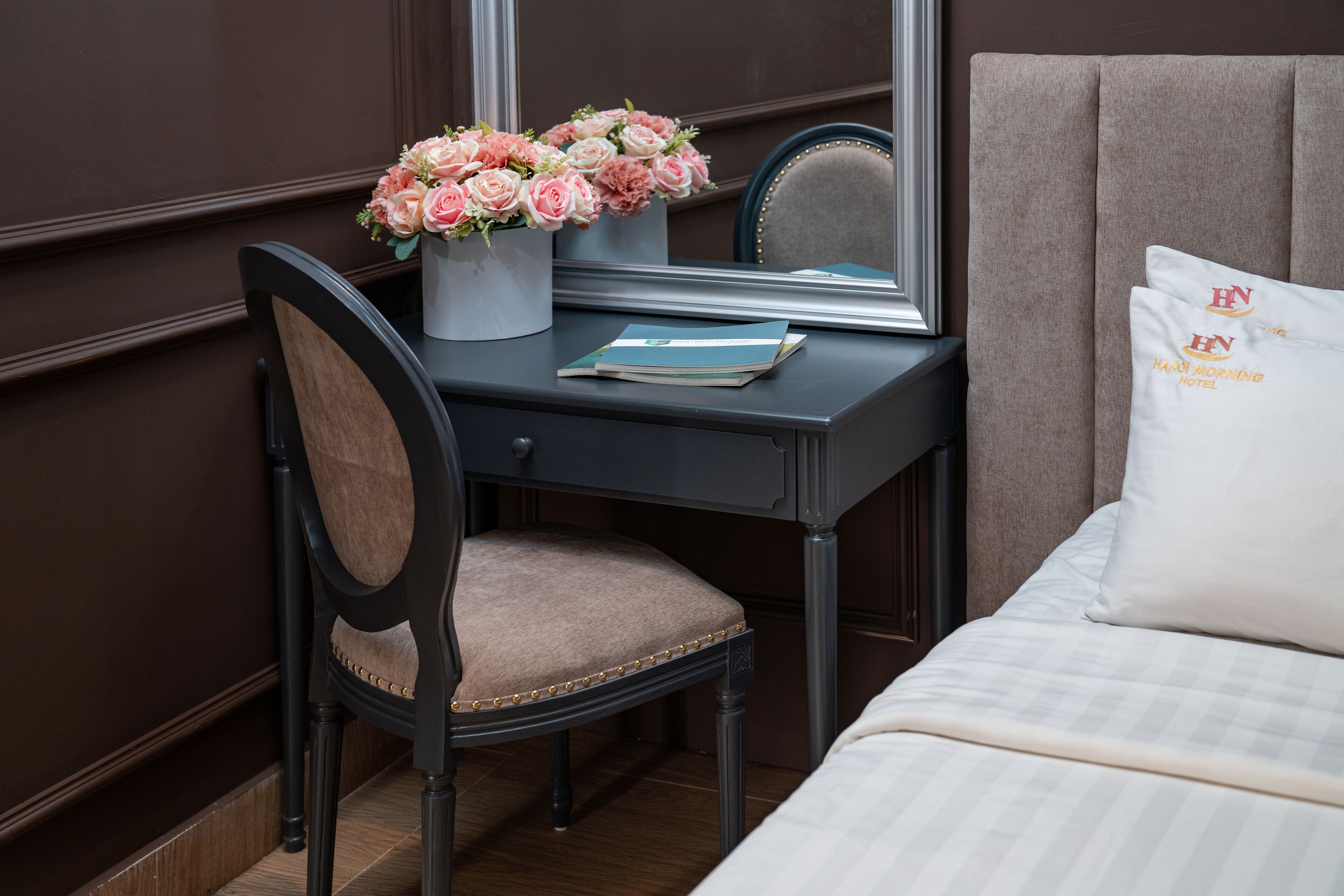 A hotel room with a bed, a desk with a chair, a mirror, and a vase of flowers.