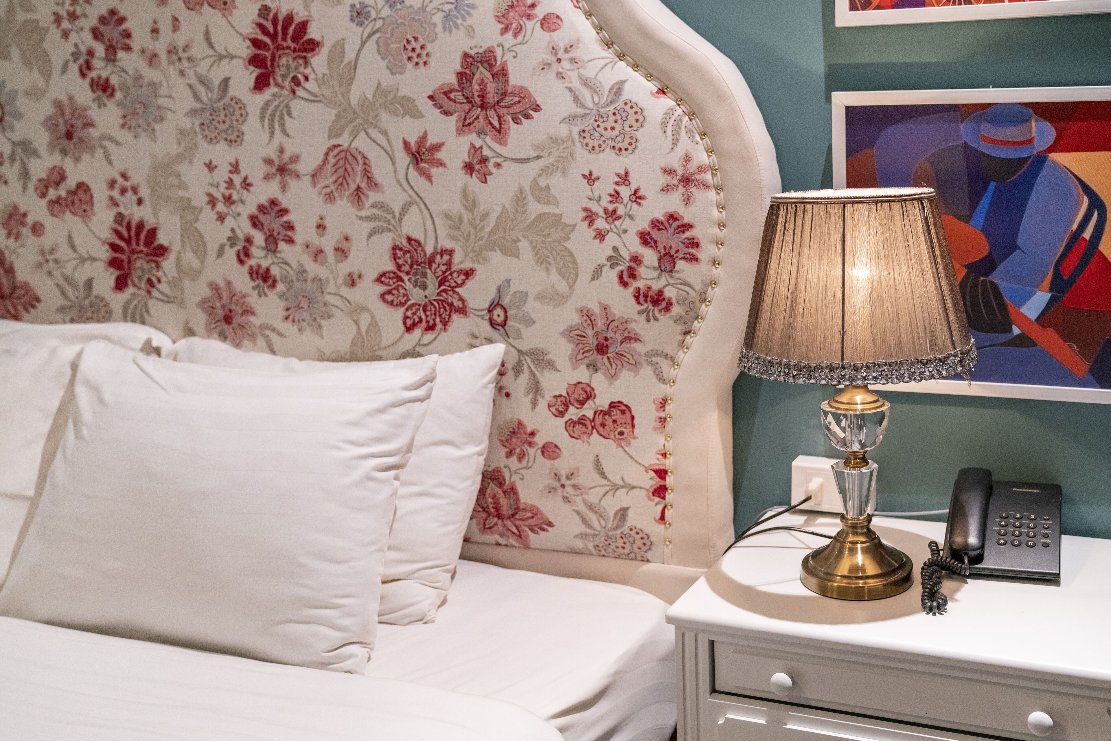 Ein ordentlich bezogenes Bett mit weißen Leinen, ein Kopfseitenverkleidung mit Blumenmuster, eine Nachttischlampe mit dekorativer Schirm und ein schwarzes Telefon auf einem weißen Nachttisch.