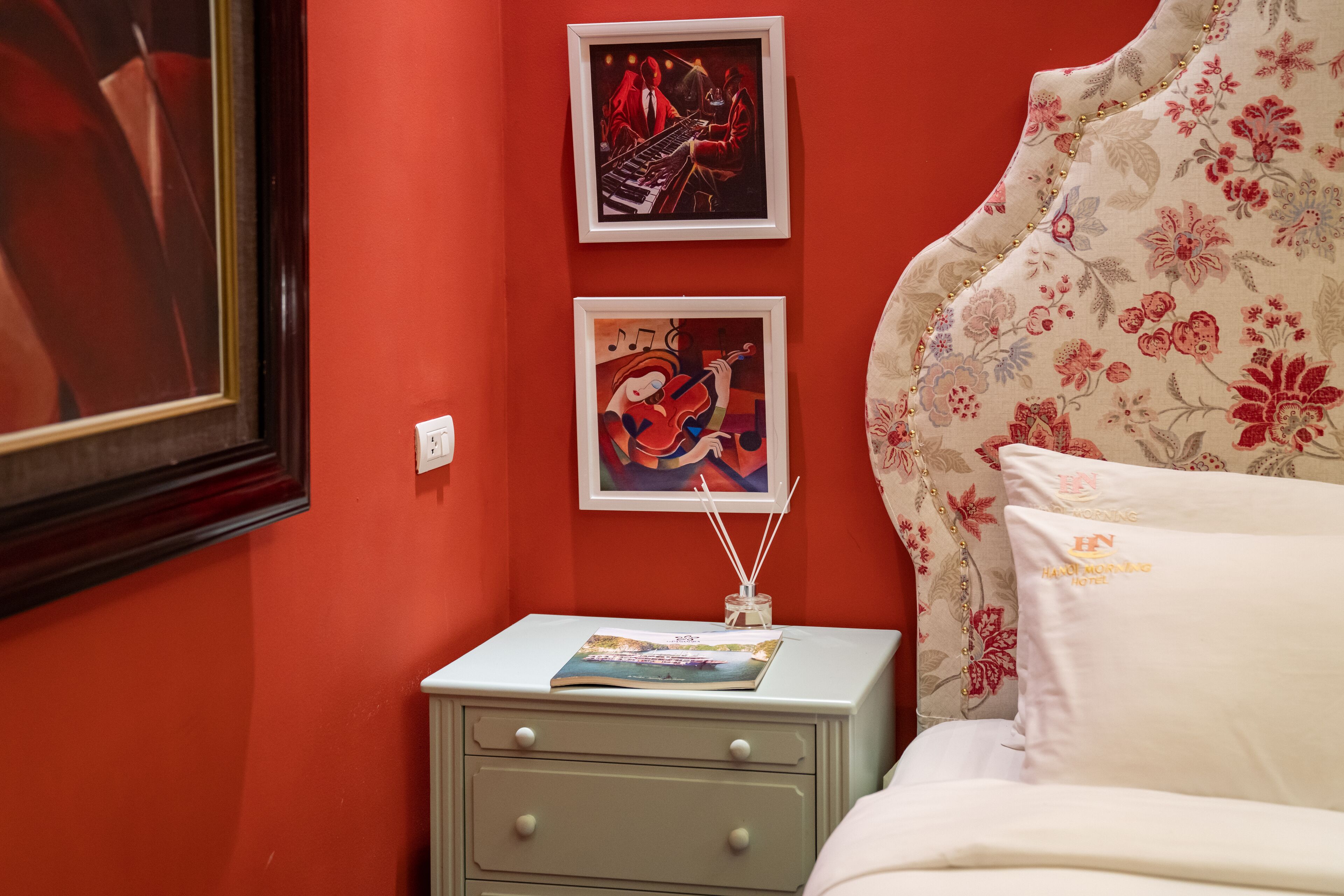 A bedroom with red walls, a nightstand with a magazine, two framed artworks, and a floral-patterned bed with a pillow labeled "HN."