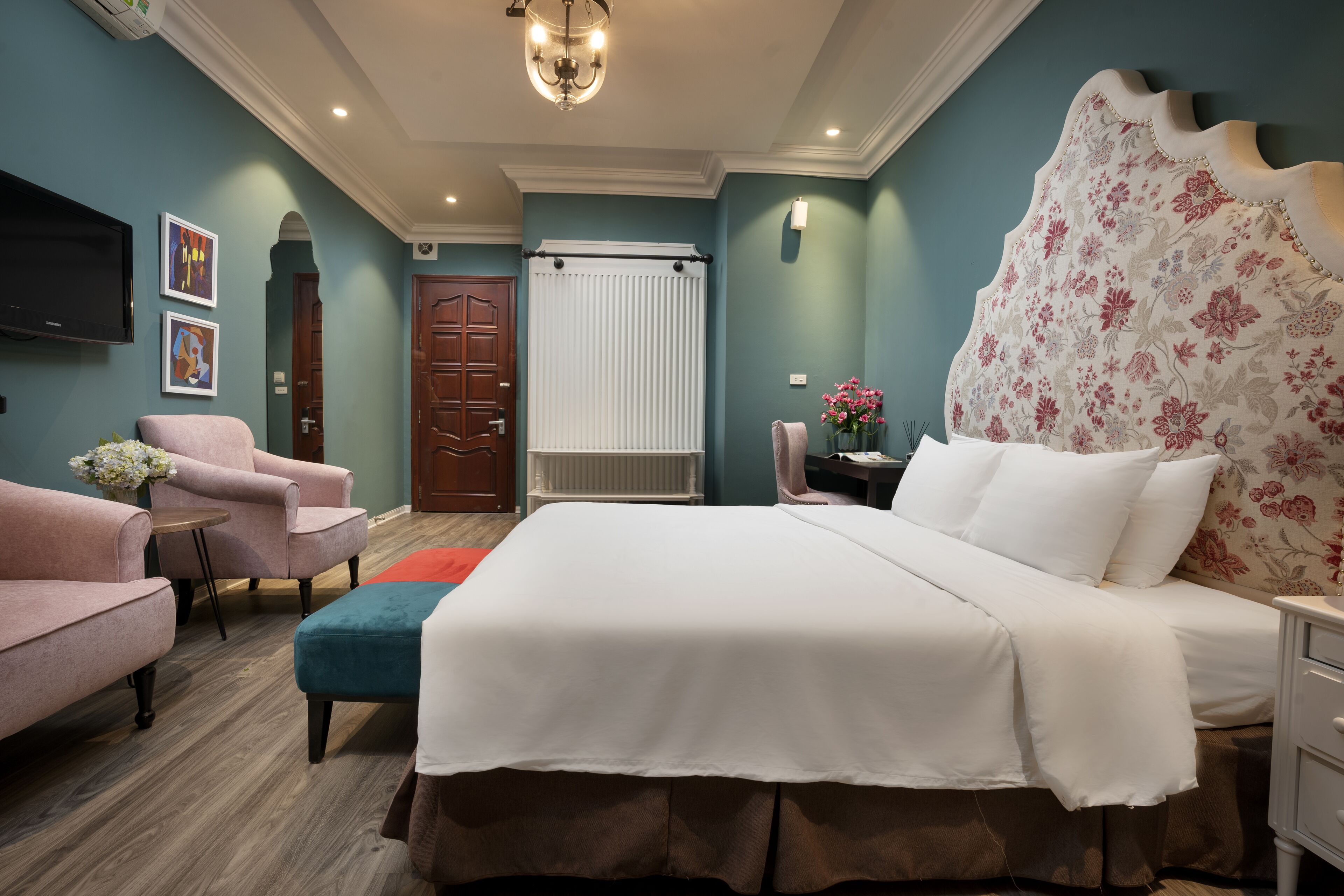A bedroom with a large bed, a chair, a small table, a nightstand, and a wall-mounted TV.