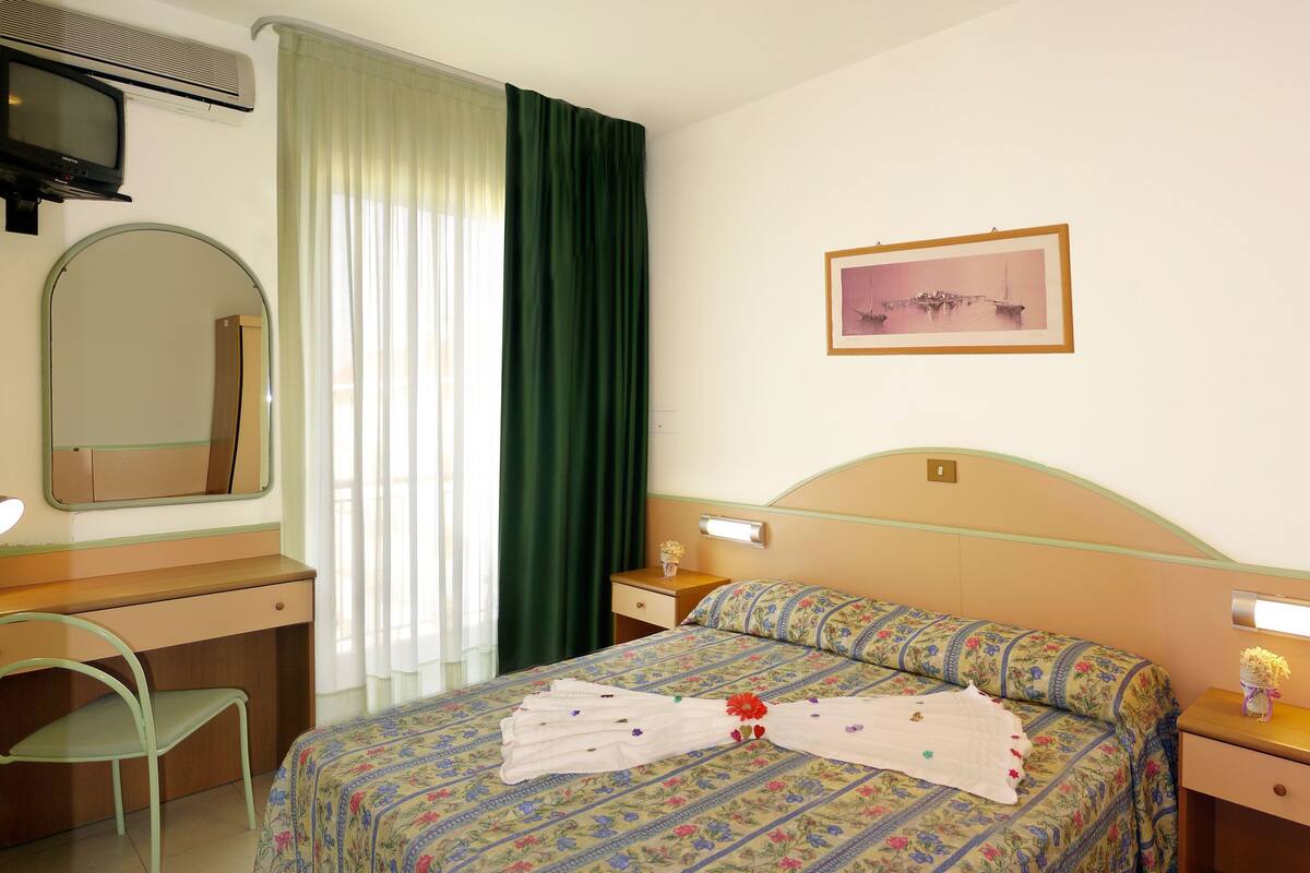 A hotel room with a bed, a desk, a chair, a mirror, a picture on the wall, and a window with curtains.