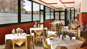 A restaurant with multiple tables set for dining, large windows offering a view of the street, and a modern ceiling design. - Hotel Beach 2 (Jesolo)