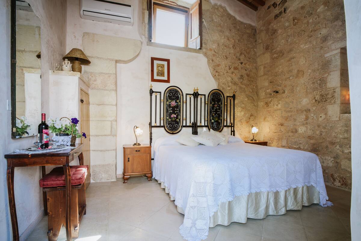 Ein Schlafzimmer mit Steinsturz, einem Bett mit weißer Bettdecke, einem Nachttisch aus Holz mit Lampe, einem Holzbüro mit Stuhl und einem Spiegel.