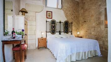 Un dormitorio con pared de piedra, una cama con cubrecama blanco, una mesita de noche de madera con lámpara, un escritorio de madera con silla y un espejo.